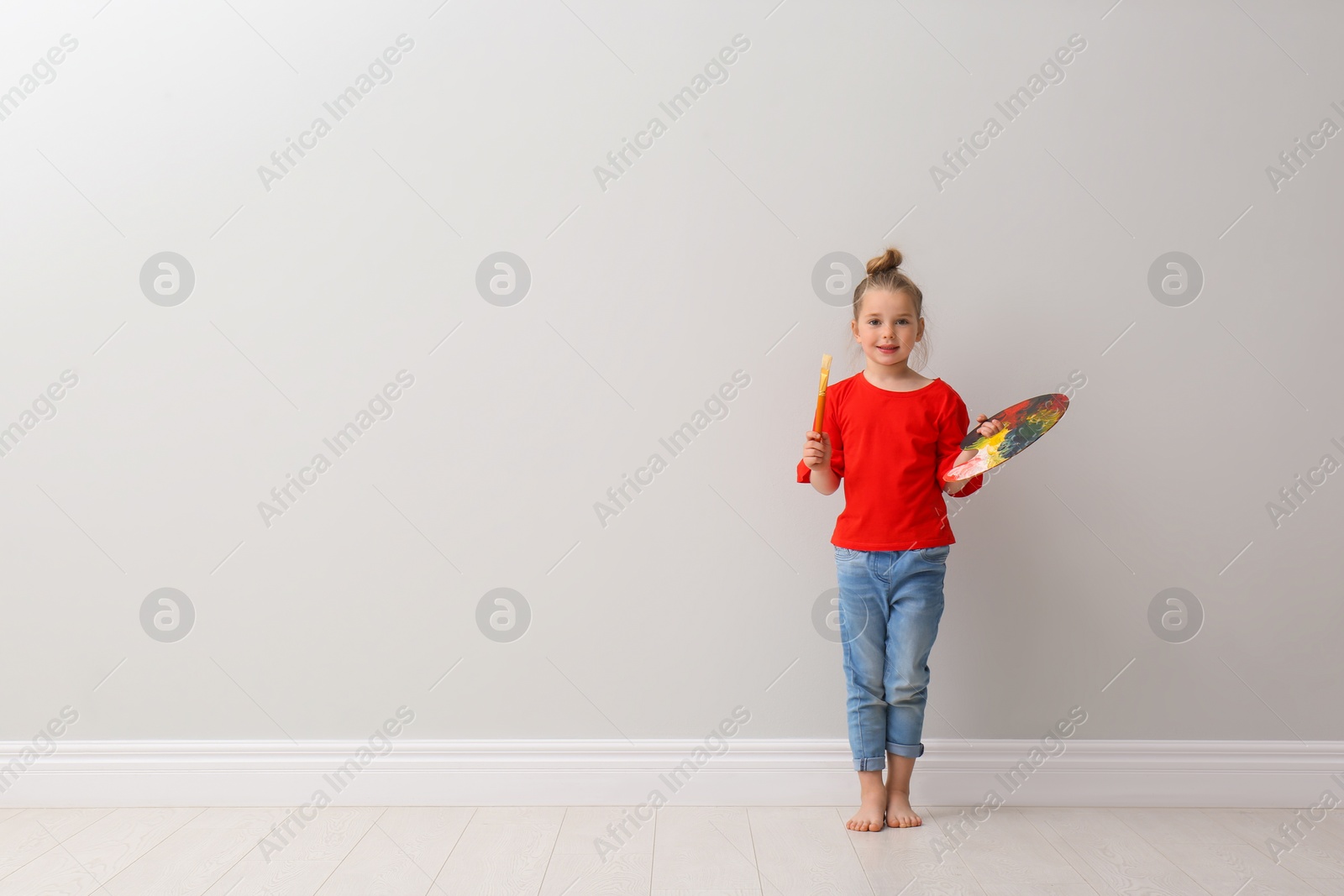 Photo of Little girl with brush and paint palette near light grey wall indoors. Space for text