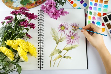 Photo of Woman painting chrysanthemums in sketchbook at white table, top view