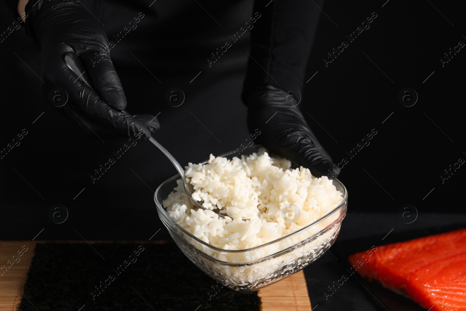 Photo of Chef in gloves taking cooked rice for sushi with spoon at dark table, closeup