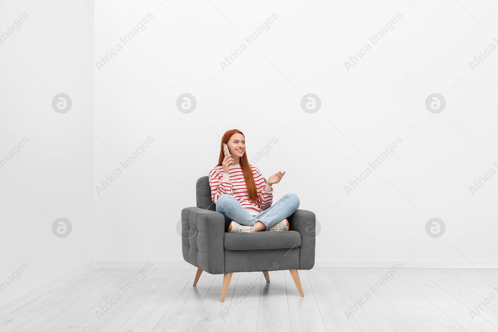 Photo of Happy young woman talking on smartphone while sitting in armchair near white wall indoors