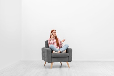 Photo of Happy young woman talking on smartphone while sitting in armchair near white wall indoors