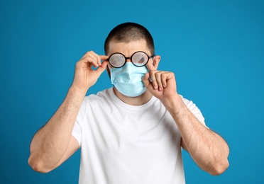 Photo of Man wiping foggy glasses caused by wearing disposable mask on blue background. Protective measure during coronavirus pandemic