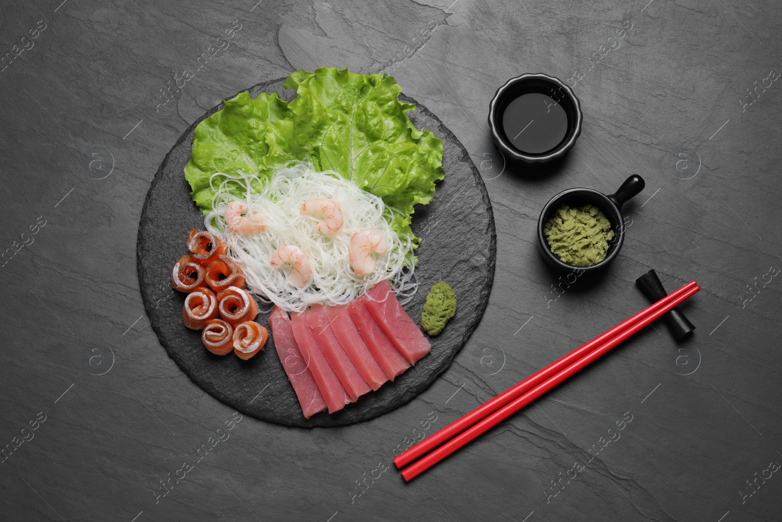 Photo of Sashimi set (raw tuna, salmon slices and shrimps) served with funchosa, lettuce, vasabi and soy sauce on dark table, flat lay