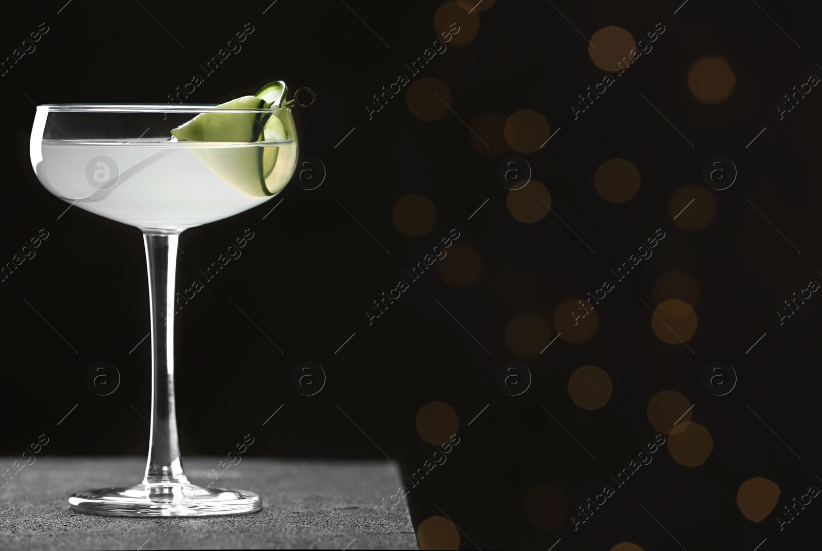 Photo of Glass of tasty cucumber martini on grey table against blurred lights, space for text