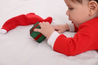 Photo of Cute baby wearing festive Christmas costume with gift box on white bedsheet, closeup