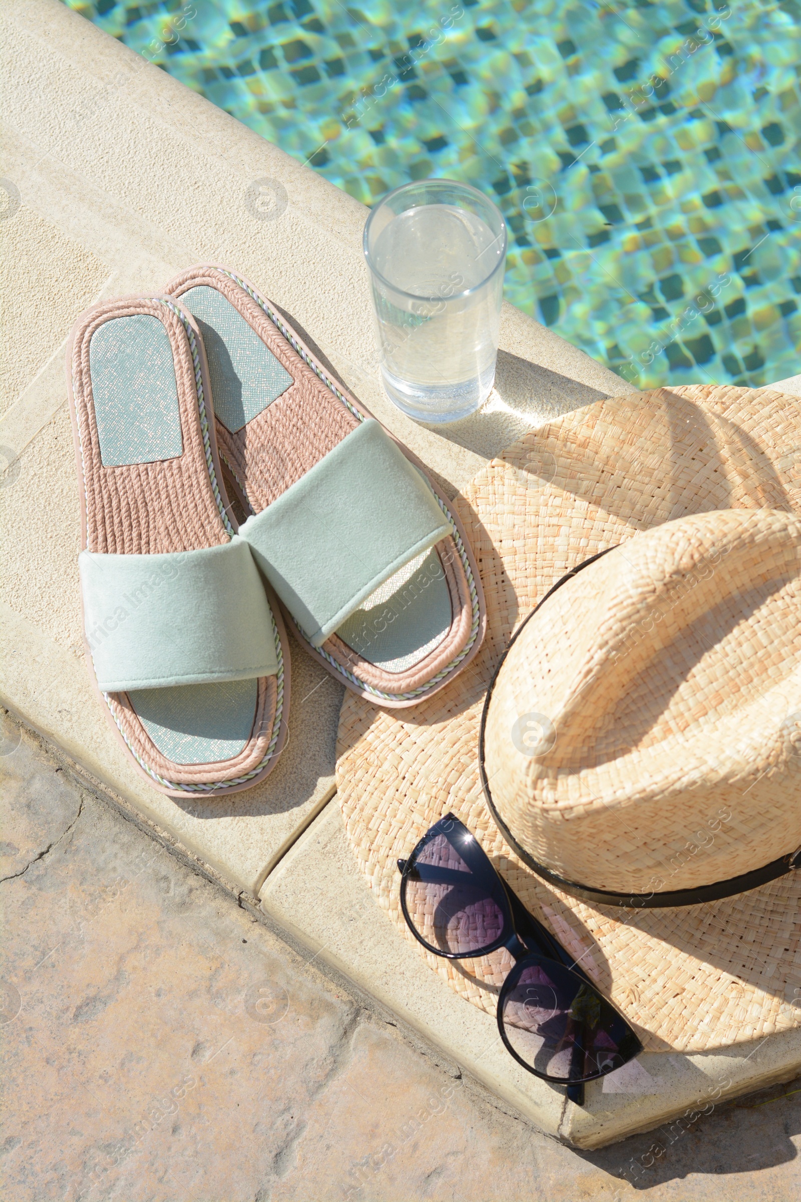 Photo of Stylish sunglasses, slippers, straw hat and glass of water at poolside on sunny day. Beach accessories