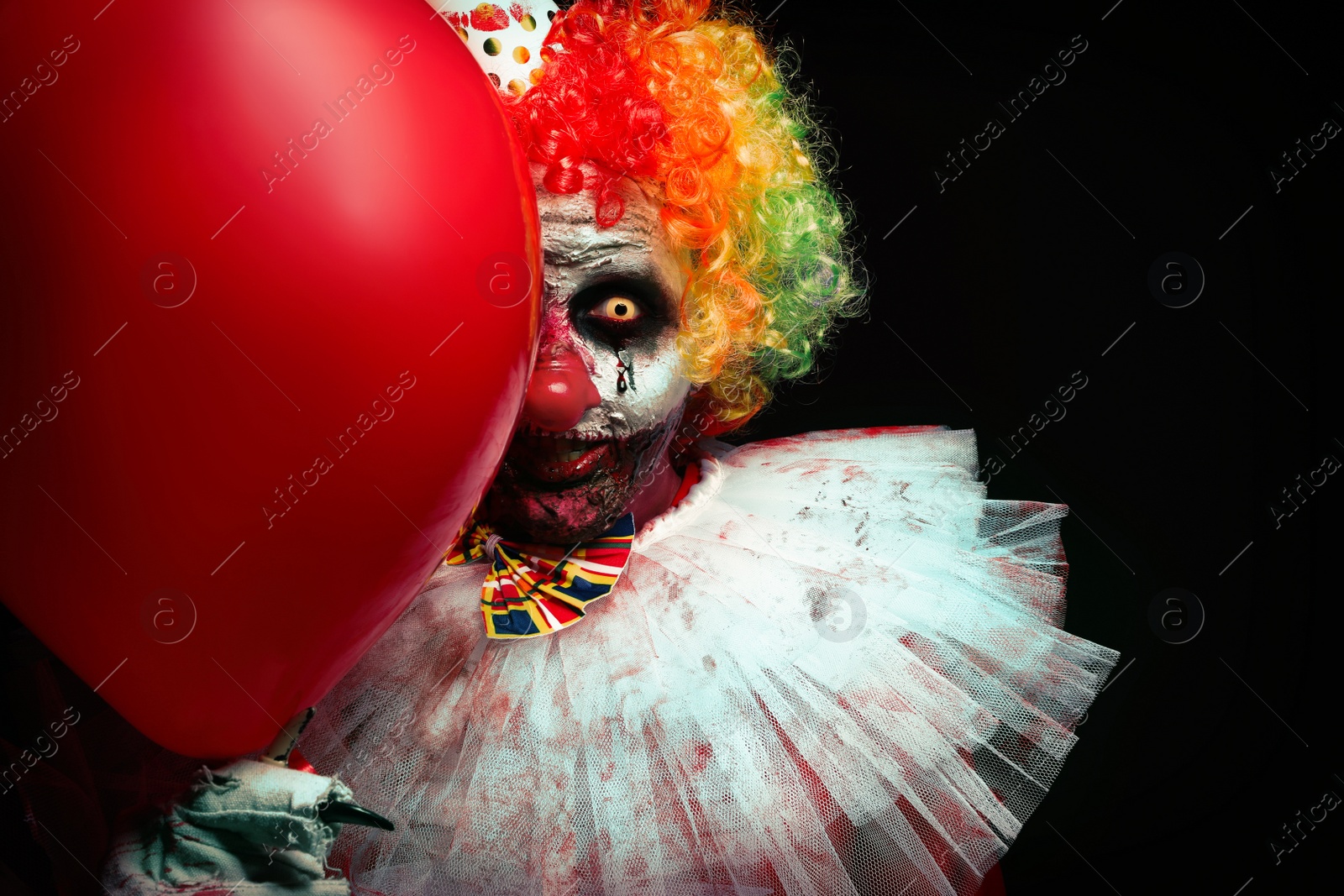 Photo of Terrifying clown with red air balloon on black background. Halloween party costume