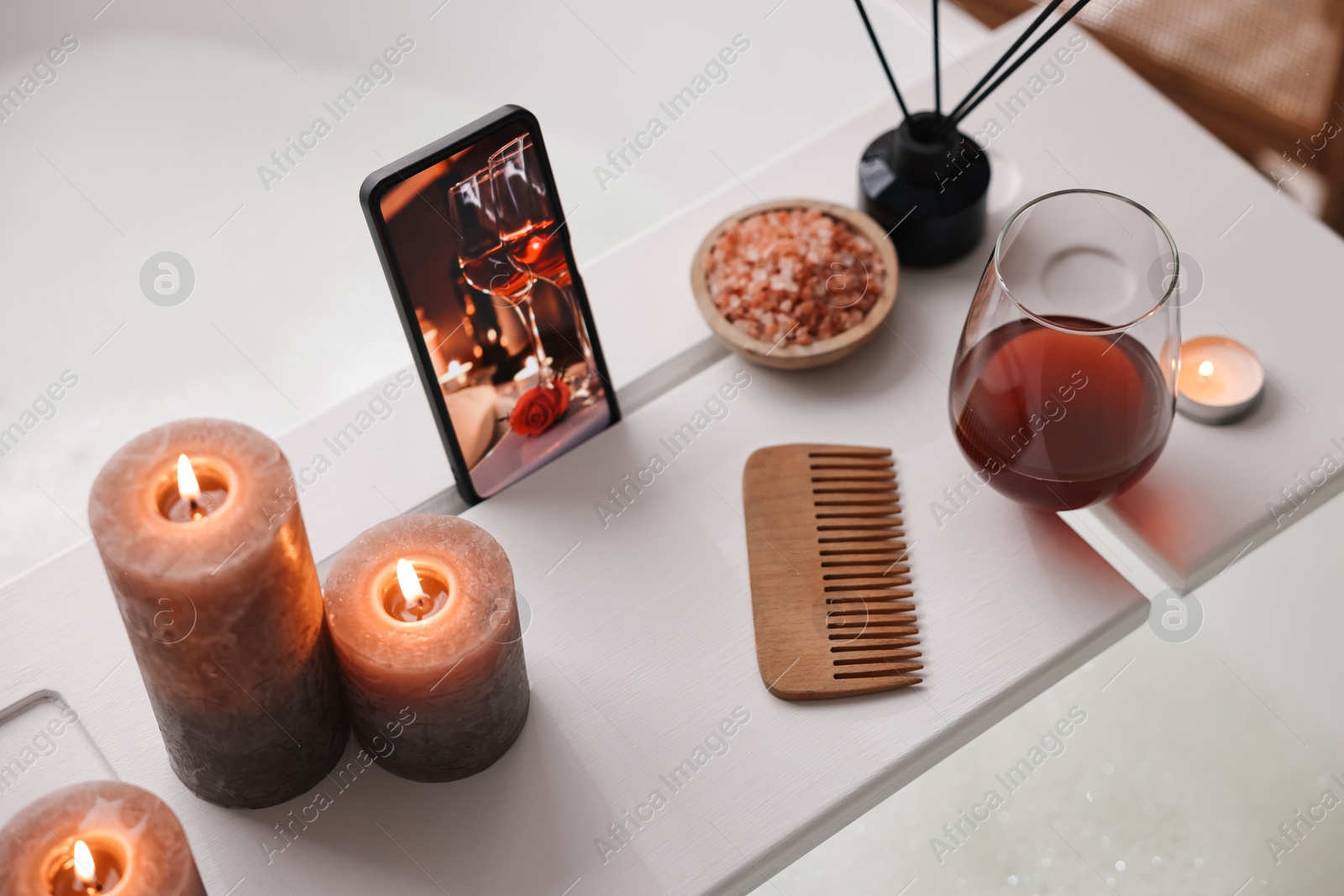 Photo of White wooden tray with smartphone, glass of wine and burning candles on bathtub