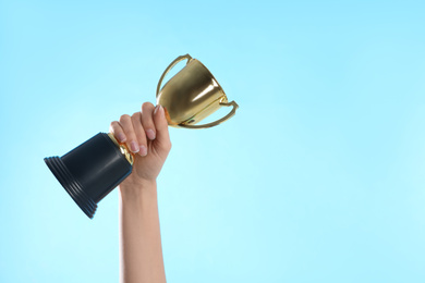 Photo of Woman holding gold trophy cup on light blue background, closeup. Space for text
