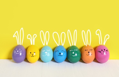 Image of Colorful eggs with drawn faces and ears as Easter bunnies on white wooden table against yellow background