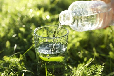 Photo of Woman pouring fresh water from bottle into glass on green grass outdoors, closeup