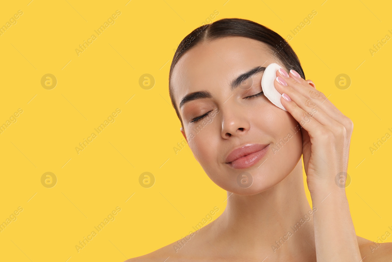 Photo of Beautiful woman removing makeup with cotton pad on yellow background. Space for text