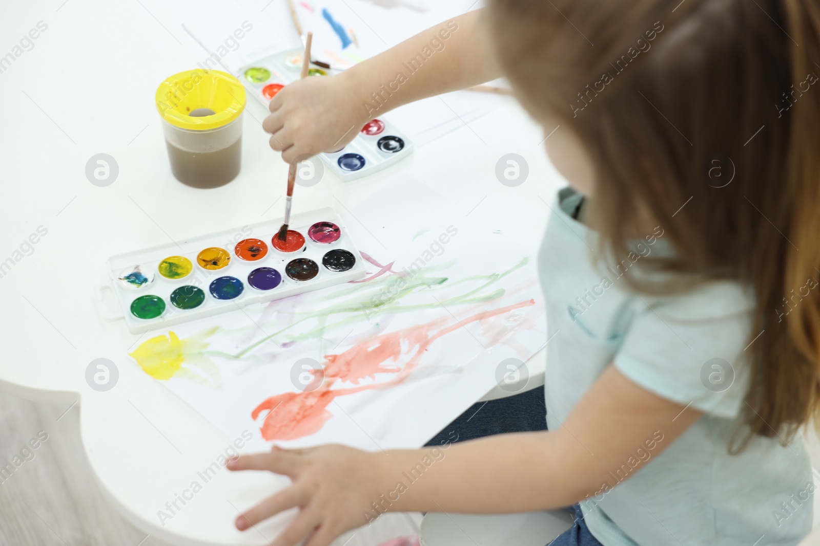 Photo of Little girl painting with brush and watercolor at white table, closeup