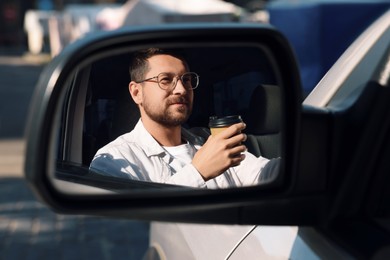 Photo of Coffee to go. Handsome man with paper cup of drink, view through car side mirror