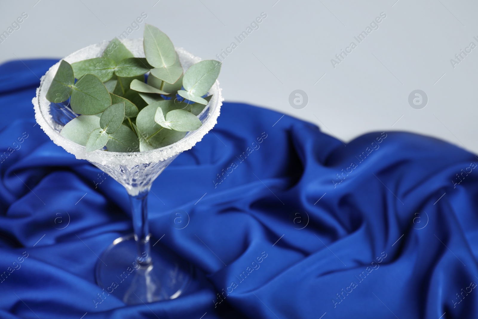 Photo of Beautiful martini glass with sugar rim and eucalyptus leaves on blue satin against light grey background. Space for text