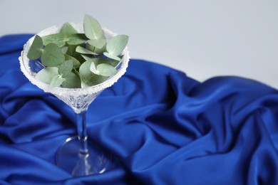 Beautiful martini glass with sugar rim and eucalyptus leaves on blue satin against light grey background. Space for text