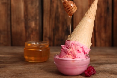 Photo of Pouring honey onto delicious pink ice cream in wafer cone on wooden table. Space for text