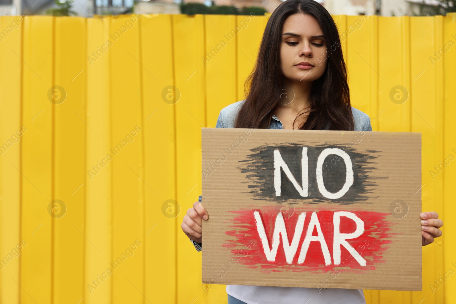 Photo of Sad woman holding poster No War near yellow fence outdoors. Space for text