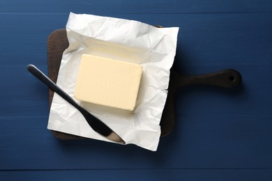 Photo of Block of tasty butter in open foil packaging and knife on blue wooden table, top view