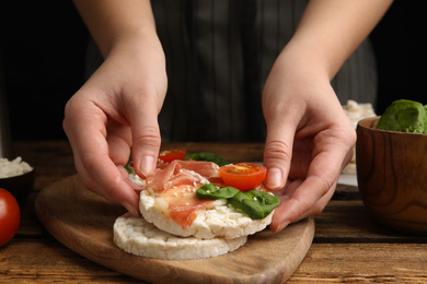 Photo of Woman holding puffed rice cake with prosciutto, tomato and basil at wooden table, closeup