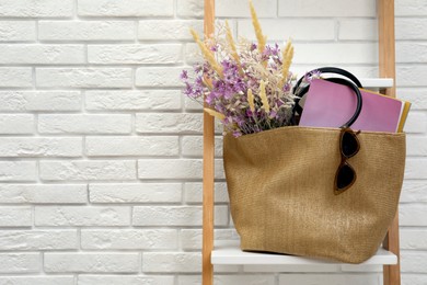 Stylish beach bag with beautiful bouquet of wildflowers, sunglasses and magazines on shelf indoors. Space for text