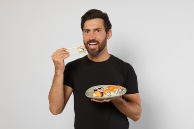 Happy man eating tasty sushi roll with chopsticks on light grey background