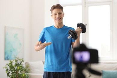 Photo of Smiling sports blogger holding dumbbell while recording fitness lesson with camera at home