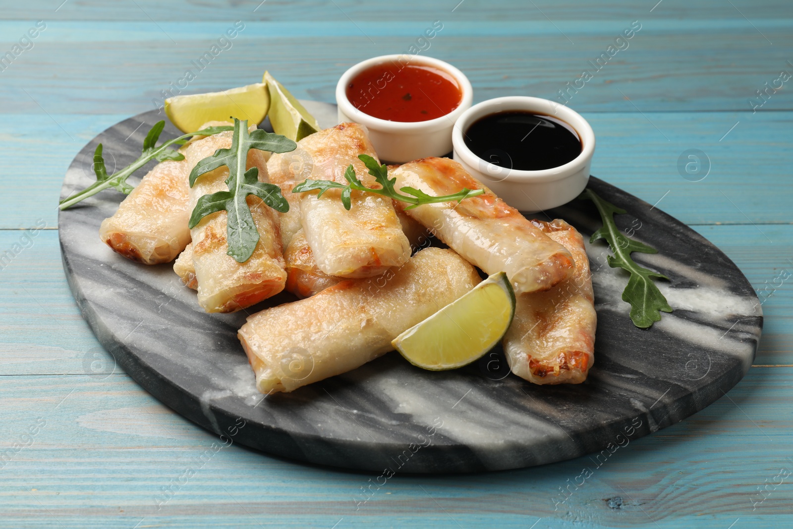 Photo of Tasty fried spring rolls, arugula, lime and sauces on light blue wooden table, closeup