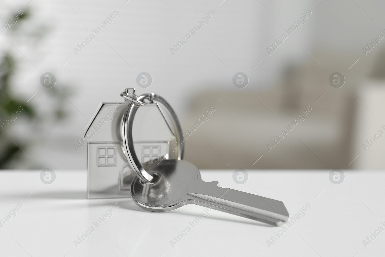 Photo of Key with keychain in shape of house on white table against blurred background, closeup. Space for text