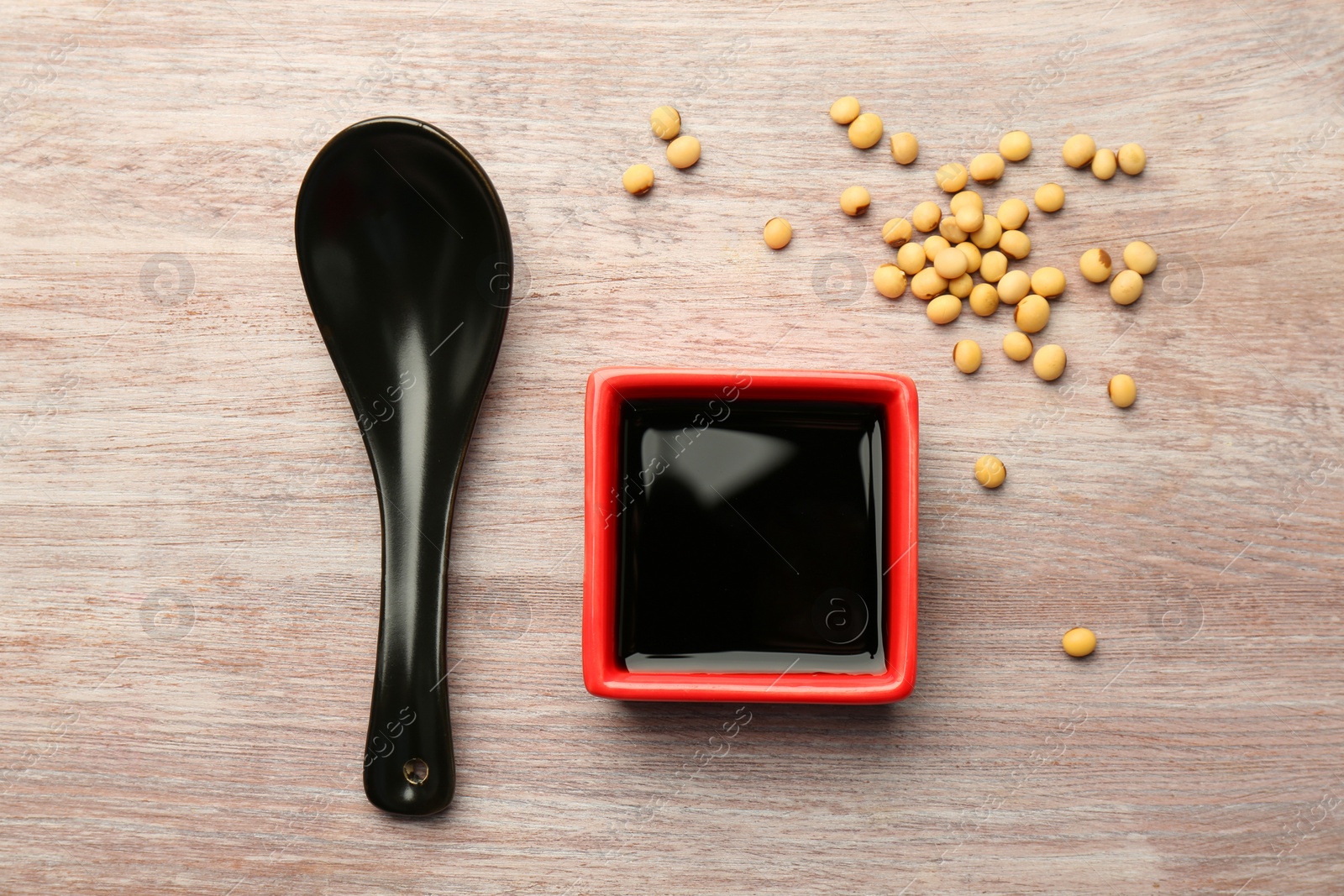 Photo of Soy sauce in bowl, soybeans and spoon on wooden table, flat lay