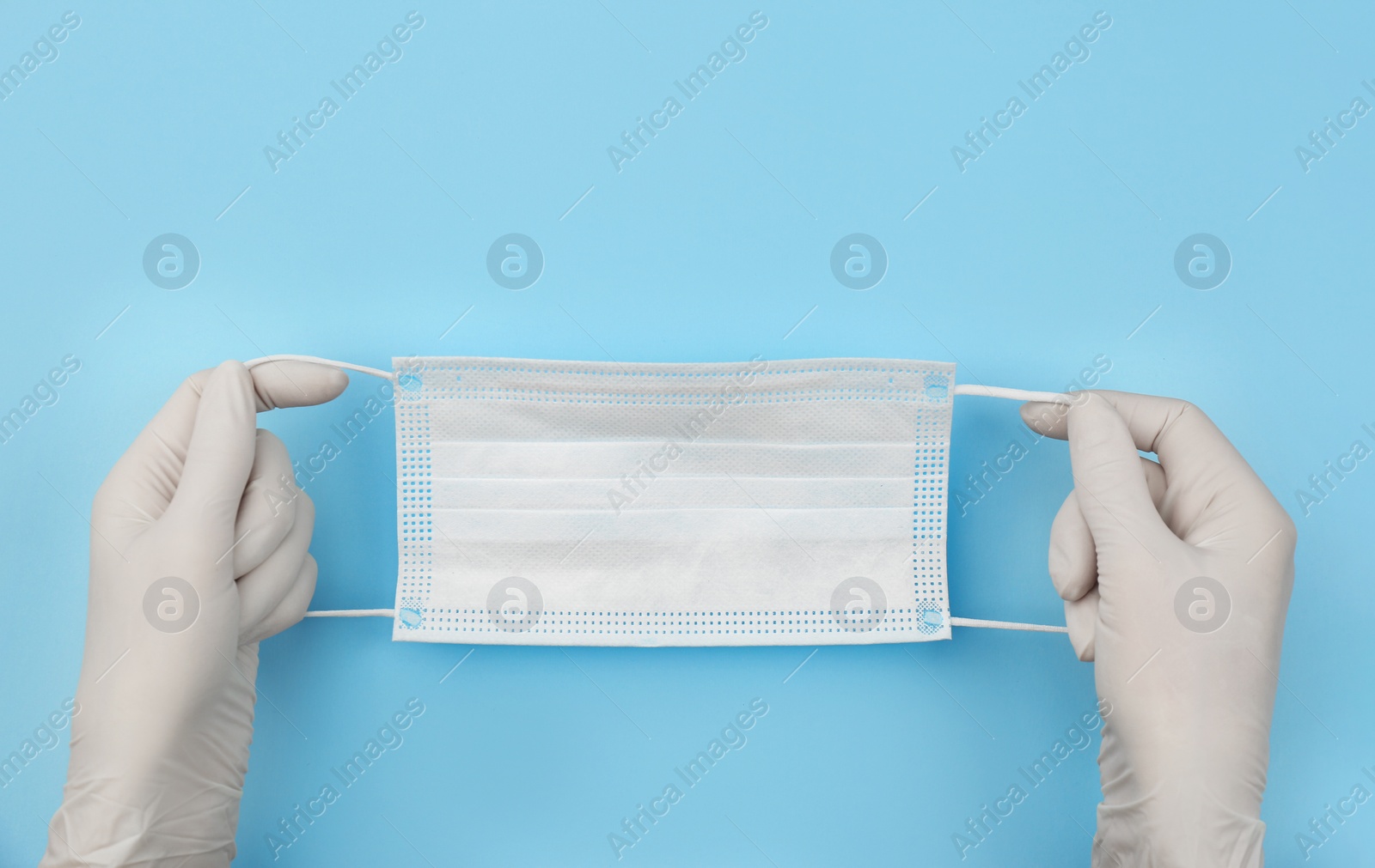 Photo of Doctor in medical gloves holding protective mask on light blue background, closeup. Space for text