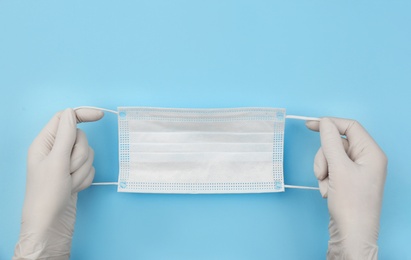 Photo of Doctor in medical gloves holding protective mask on light blue background, closeup. Space for text