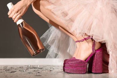 Photo of Stylish party. Woman wearing pink high heeled shoes with platform and square toes holding bottle of wine indoors, closeup