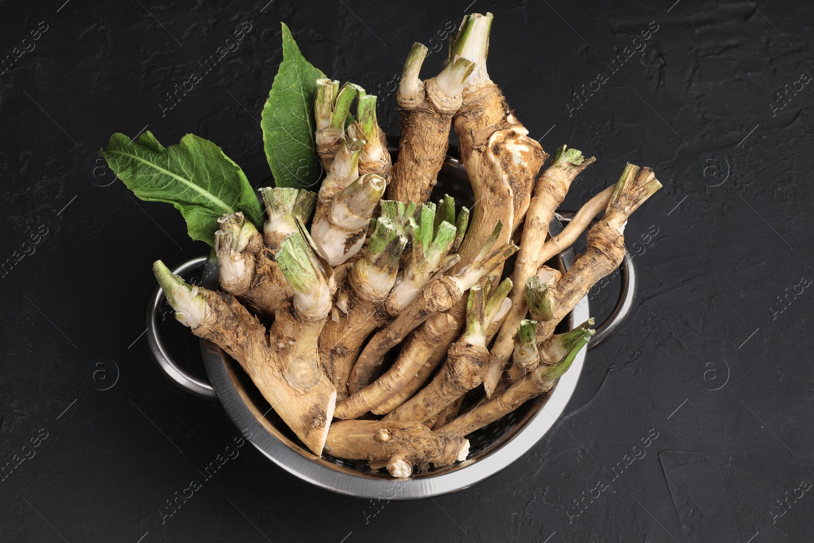 Photo of Fresh horseradish roots and leaves in colander on black textured table, above view