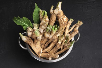 Photo of Fresh horseradish roots and leaves in colander on black textured table, above view