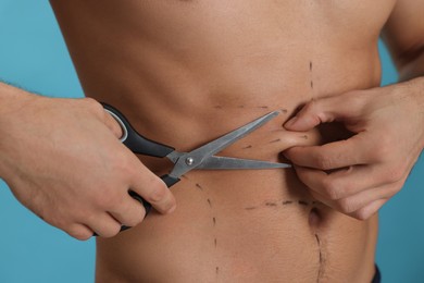 Fit man with scissors and marks on body against light blue background, closeup. Weight loss surgery