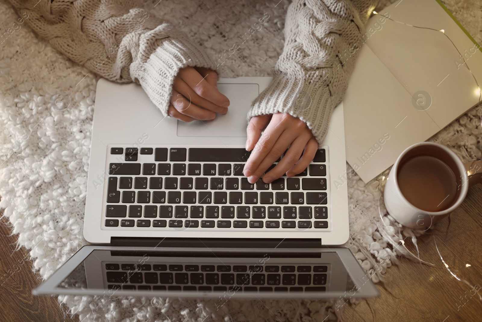 Photo of Woman with cup of hot beverage using laptop at home in winter evening, closeup