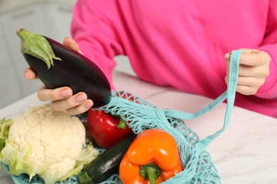 Photo of Woman taking eggplant out from string bag at light marble table, closeup