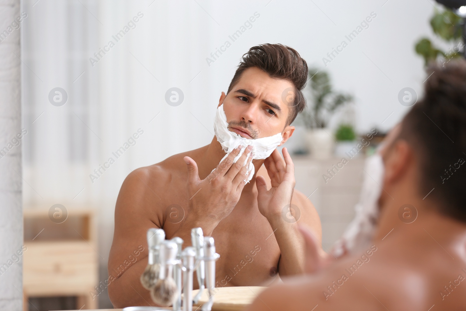 Photo of Young man applying shaving foam near mirror in bathroom