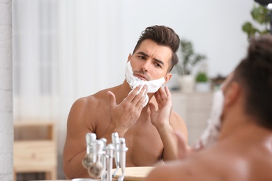 Young man applying shaving foam near mirror in bathroom