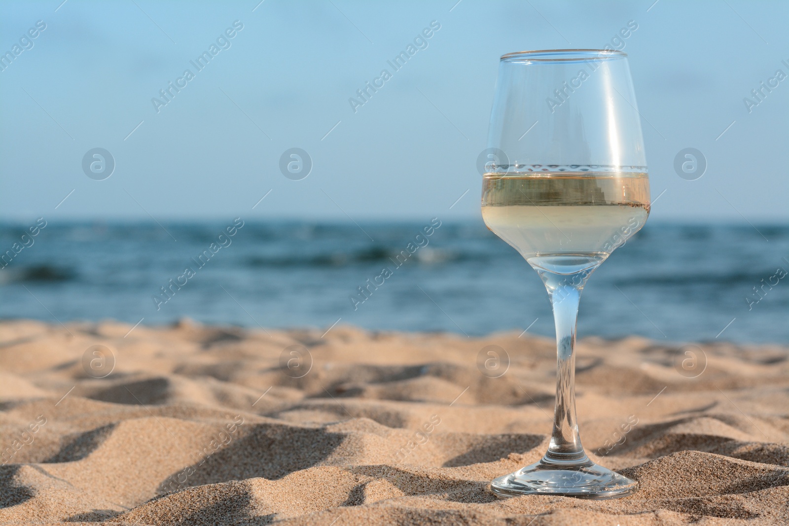 Photo of Glass of tasty wine on sand near sea, space for text