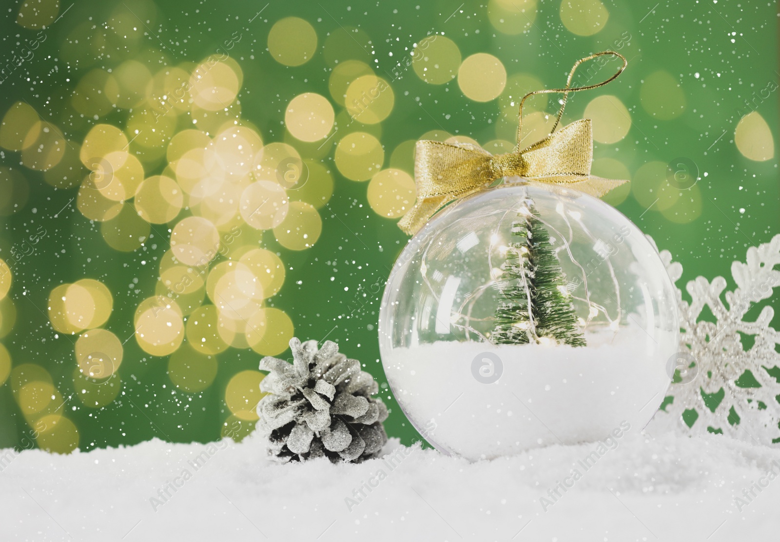 Image of Beautiful transparent Christmas ornament with small fir tree and fairy lights on snow against green background, bokeh effect. Space for text