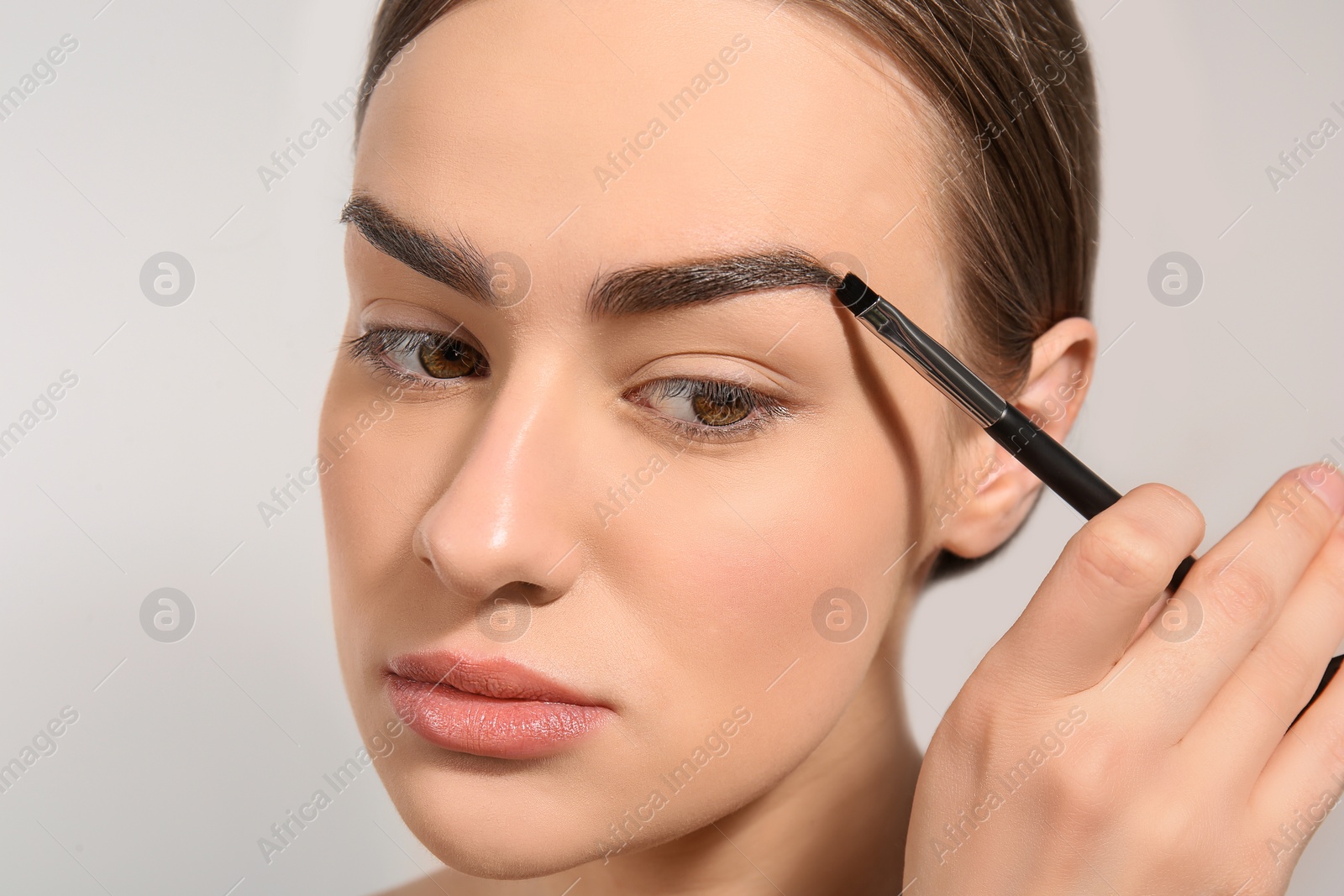 Photo of Young woman correcting shape of eyebrow with brush on light background