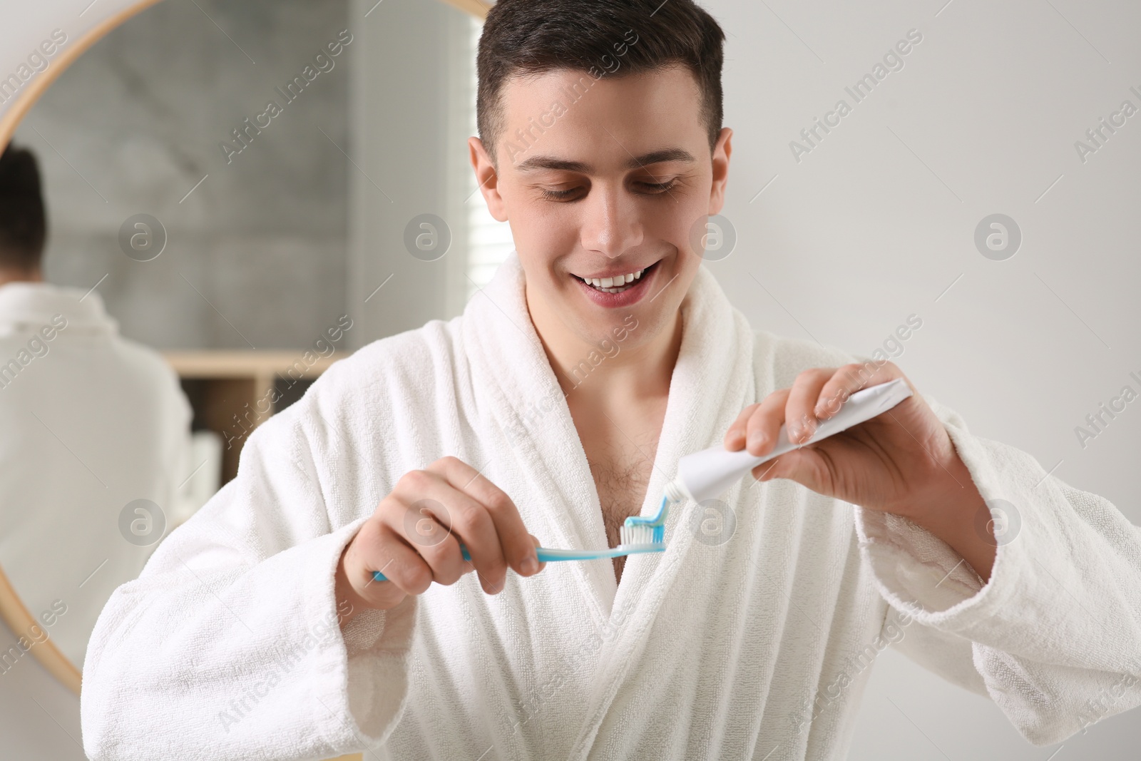 Photo of Happy man squeezing toothpaste from tube onto toothbrush in bathroom