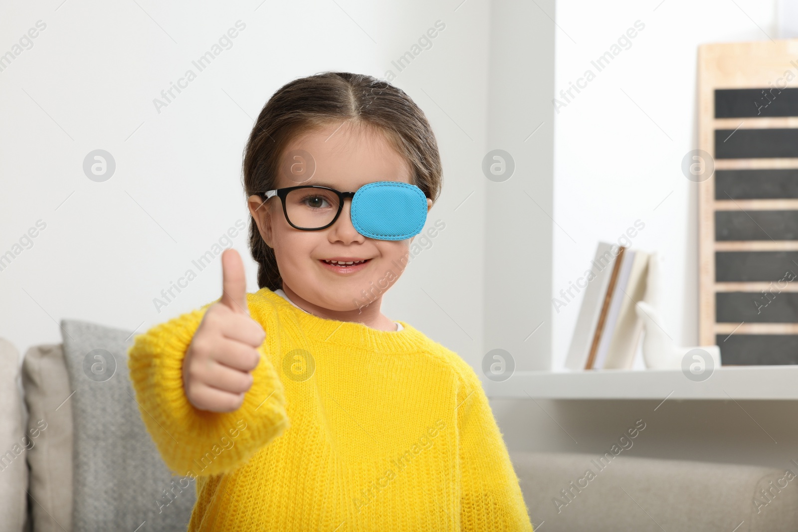 Photo of Happy girl with nozzle on glasses for treatment of strabismus showing thumbs up in room. Space for text