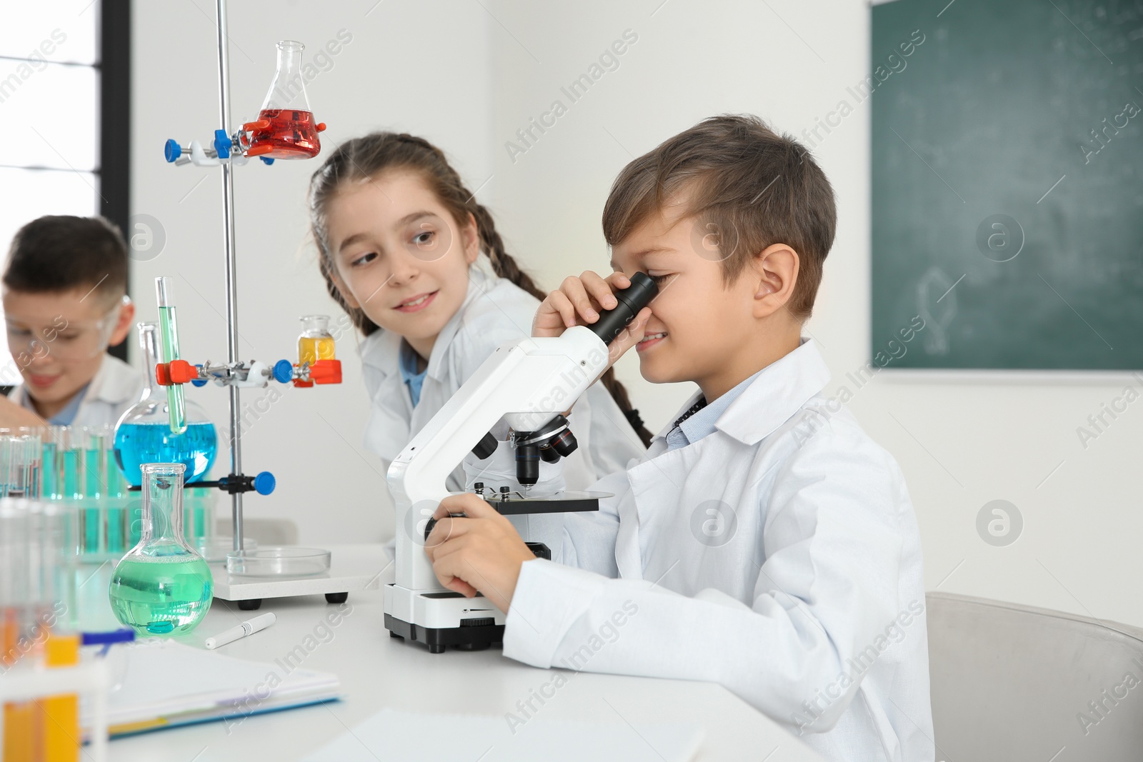 Photo of Schoolboy looking through microscope and his classmates at chemistry lesson