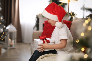 Cute little boy holding gift box on sofa in room decorated for Christmas