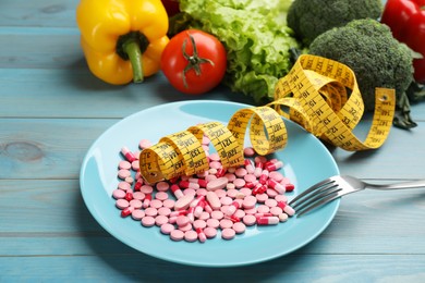 Photo of Plate with weight loss pills, measuring tape and fork near pile of vegetables on light blue wooden table