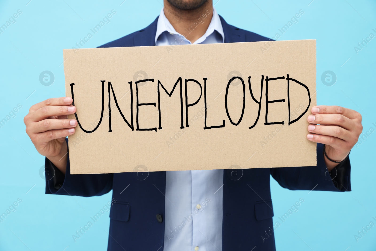Photo of Young man holding sign with word Unemployed on light blue background, closeup