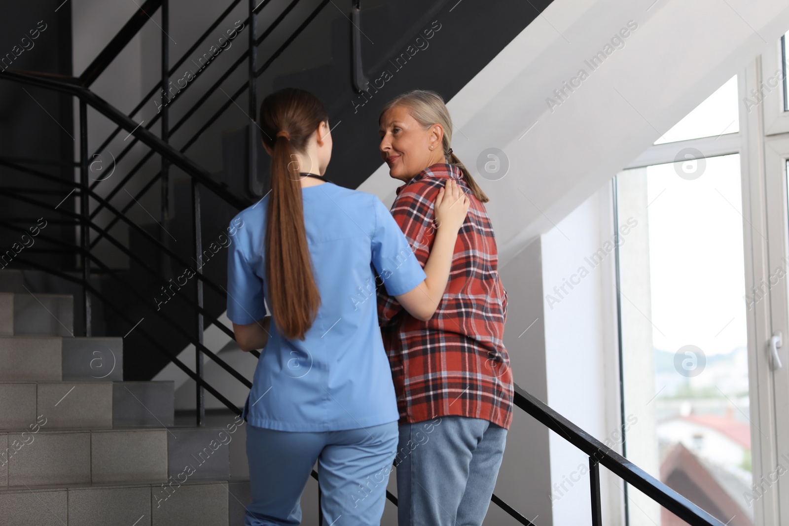 Photo of Young healthcare worker assisting senior woman on stairs indoors, low angle view